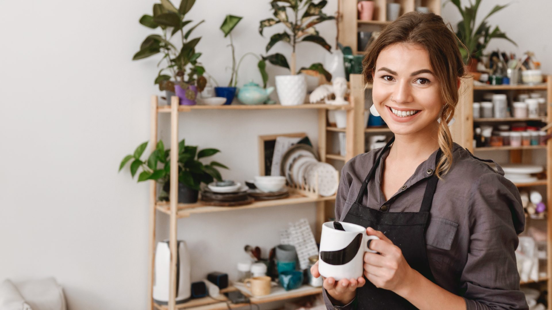 Artesã sorrindo em seu ateliê, segurando uma caneca feita à mão